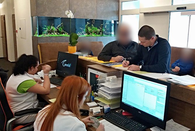 Pacjenci przy rejestracji w klinice, obok personel przy komputerach